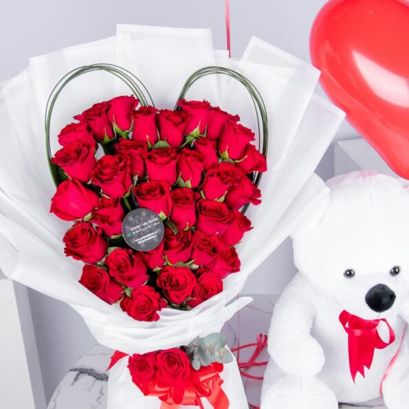 A close up view of Red Roses arranged in a Heart Shape with Steel Grass, heart shaped balloons and a White Teddy Bear