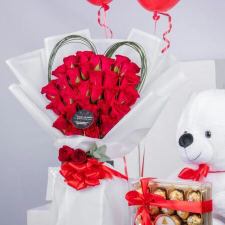 A bouquet of red roses, a box of chocolates, a teddy bear, & two heart-shaped balloons