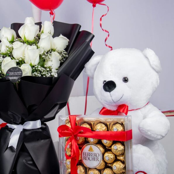 A bouquet of white roses, a box of chocolates, a teddy bear, & two heart-shaped balloons
