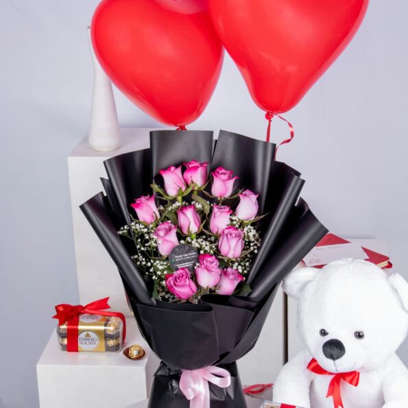 A bouquet of pink roses, a box of chocolates, a teddy bear, & two heart-shaped balloons